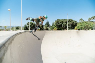 Delikanlı, beton bir kaykay kasesinde scooterıyla bir numara yapıyor.