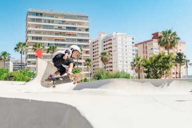 Şehir kaykay parkında kaykay antrenmanı yapan, koruyucu kıyafetler giyen ve güneşli bir günde eğlenen bir çocuk.