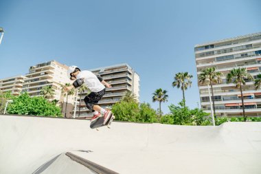 Şehir kaykay parkında kaykay antrenmanı yapan, koruyucu kıyafetler giyen ve numaralar yapan bir çocuk.