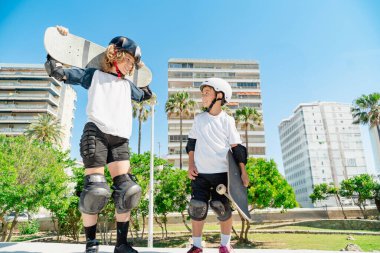 Kasklı ve koruyucu teçhizatlı iki neşeli kaykaycı bir kaykay parkında birbirlerine gülümsüyorlar.