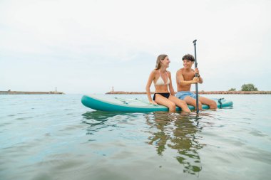 Genç bir çift kürek tahtasında oturuyor, suda rahatlatıcı bir günün tadını çıkarıyorlar.