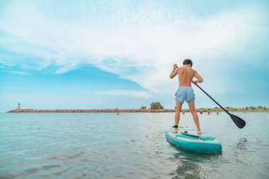 Sahile yakın denizde sörf yaparak yaz tatilinin tadını çıkarmaya çalışan genç bir adam.