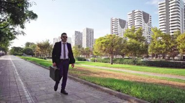 Takım elbiseli ve güneş gözlüklü bir işadamı modern bir şehir parkında evrak çantasıyla yürüyor.