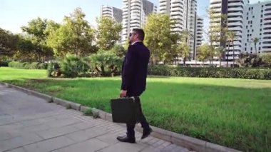 Takım elbiseli genç bir girişimci şehir parkında ofis binaları olan bir kaldırımda yürüyor.