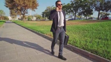 Elinde evrak çantası olan şık bir işadamı telefonda konuşurken parkta yürüyor.