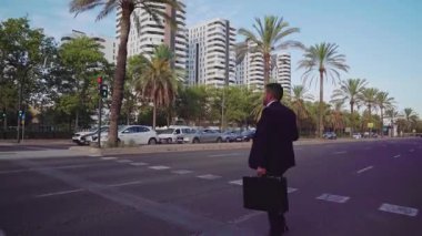 Takım elbiseli bir girişimci modern bir şehirde palmiye ağaçlarıyla yaya geçidinde karşıdan karşıya geçiyor.
