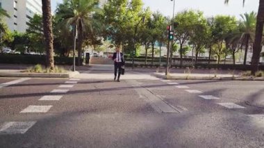 Orta yaşlı takım elbiseli bir yönetici elinde çanta ve kahveyle kameraya doğru yürüyor.