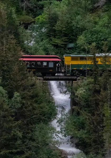 Train conductor is standing outside while the colorful train passes over a waterfall.  White Pass, Alaska.