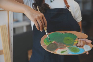 Bir sehpanın önünde duran ve fırçayı renk paletine batıran kadın sanatçının kırpılmış görüntüsü.