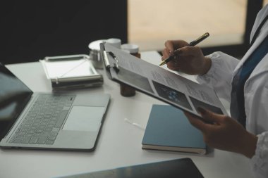 Ciddi bir kadın doktor dizüstü bilgisayar kullanıyor ve tıp dergisine notlar yazıyor. Beyaz önlüklü ve steteskoplu genç kadın doktor iş yerindeki bilgisayarda çalışıyor..