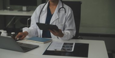 Ciddi bir kadın doktor dizüstü bilgisayar kullanıyor ve tıp dergisine notlar yazıyor. Beyaz önlüklü ve steteskoplu genç kadın doktor iş yerindeki bilgisayarda çalışıyor..