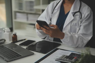 Ciddi bir kadın doktor dizüstü bilgisayar kullanıyor ve tıp dergisine notlar yazıyor. Beyaz önlüklü ve steteskoplu genç kadın doktor iş yerindeki bilgisayarda çalışıyor..