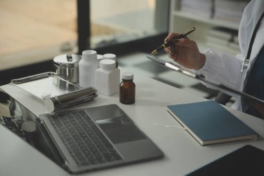 Ciddi bir kadın doktor dizüstü bilgisayar kullanıyor ve tıp dergisine notlar yazıyor. Beyaz önlüklü ve steteskoplu genç kadın doktor iş yerindeki bilgisayarda çalışıyor..