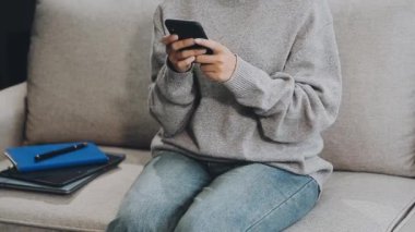 Genç Asyalı kadın evdeki kanepede akıllı telefon kullanıyor.