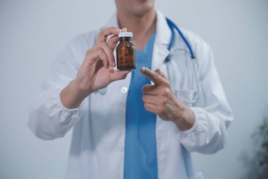 Genç Asyalı erkek doktorun portresi gülümsüyor, ilaç şişesini gösteriyor ve hastanede dostça ve nazik bir şekilde dururken kendinden emin bir şekilde bakıyor. Sağlık ve sağlık sigortası kavramı.