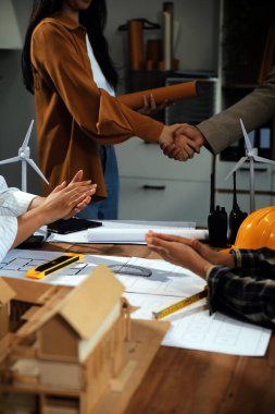 İnşaat ekibi yeni proje planını başlatmak için ofis merkezindeki sarı kaskın arkasında el sıkışıyor ve inşaat projeleriyle ilgili danışmanlık yapıyor. Güneş ışığında