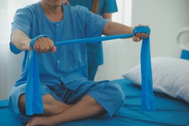 Hasta esnek bir egzersiz bandıyla esneme egzersizi yapıyor ve klinikte yardımcı olmak için fizyoterapist eli kullanıyor..