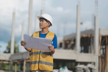 Asyalı mühendis ya da mimar ellerinde inşaat alanında beyaz kasklı evrak planları tutuyor. Modern binada duruyor..