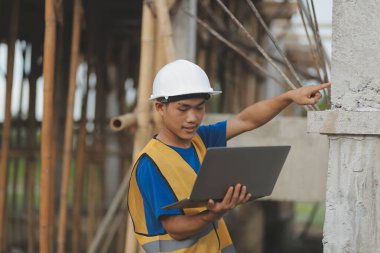 Genç yakışıklı Asyalı inşaat mühendisi elinde tabletle ileriye bakıyor ve düşünüyor, inşaat geçmişini planlıyor. Evrim inşa kavramı.