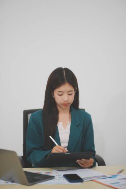 Güzel Asyalı iş kadını, ofise yerleştirilmiş evraklarla birlikte tablet ve dizüstü bilgisayar kullanıyor..