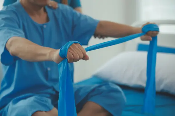 Hasta esnek bir egzersiz bandıyla esneme egzersizi yapıyor ve klinikte yardımcı olmak için fizyoterapist eli kullanıyor..