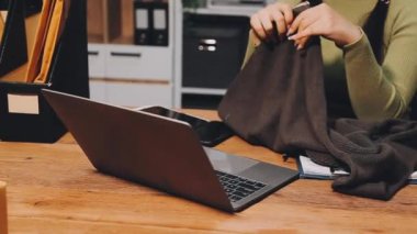 Kadın elleri dizüstü bilgisayarı tutuyor ve mutfaktaki tahta masada otururken akıllı telefon kullanıyor.