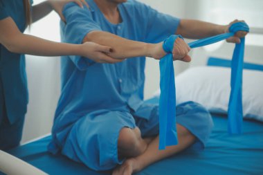 Hasta esnek bir egzersiz bandıyla esneme egzersizi yapıyor ve klinikte yardımcı olmak için fizyoterapist eli kullanıyor..