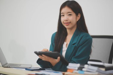 Güzel Asyalı iş kadını, ofise yerleştirilmiş evraklarla birlikte tablet ve dizüstü bilgisayar kullanıyor..