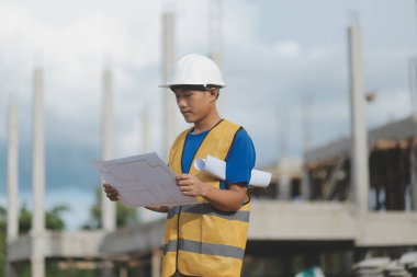 Asyalı mühendis ya da mimar ellerinde inşaat alanında beyaz kasklı evrak planları tutuyor. Modern binada duruyor..