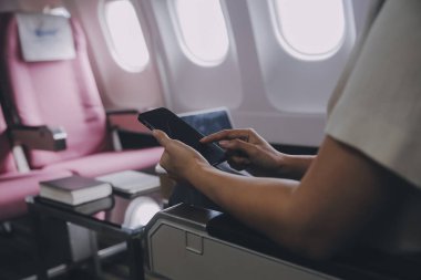 Pencerenin yanında uçakta oturan ve sosyal ağlardan haber okuyan veya akıllı telefonda seyahat uygulamalarını kullanan Asyalı kadın yolcu