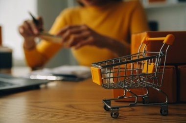 Akıllı telefon tutan genç bir kadın, ödeme başarısını gösteren bir tablet ve sarı paket kutusu olan kredi kartını ofiste online alışveriş kavramı olarak gösteriyor.