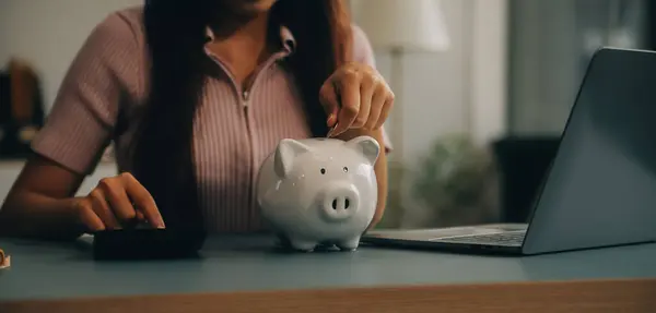 Young Asian woman saving for retirement Saving money through a piggy bank and taking notes on notebook, savings concept.
