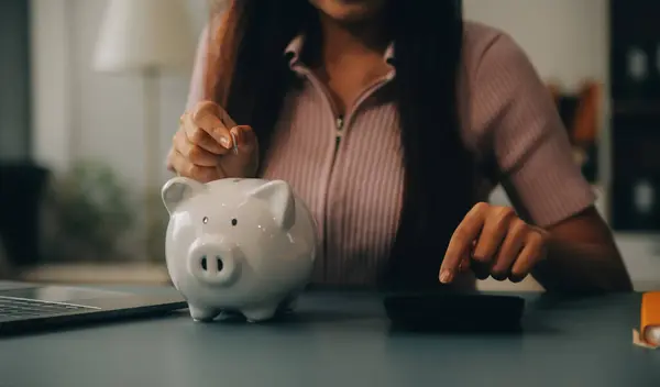 Young Asian woman saving for retirement Saving money through a piggy bank and taking notes on notebook, savings concept.