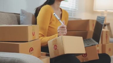 Odasında bir sürü karton kutu olan Asyalı bir kadın. Çevrimiçi ürünler satan genç kadın girişimci, indirme ve çevrimiçi iş aracı 