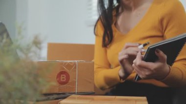Asyalı bir kadın bir odada sayısız karton kutularla çevrili. Çevrimiçi satış, indirme ve e-ticaret yapan genç bir girişimci olarak rolünü yansıtıyor.