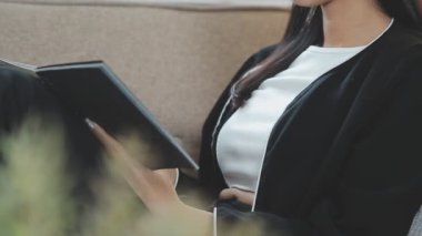 Güzel Asyalı kadın kanepede oturuyor ve siyah defterden notlar okuyor.