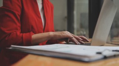 Kırmızı elbiseli Asyalı kadın modern ofisteki dizüstü bilgisayarında çalışıyor.