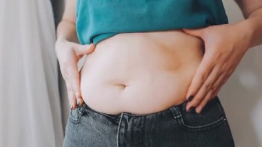 Genç bir kadın şişman karnına elleriyle dokunuyor, kilolu olma kavramını temsil ediyor.