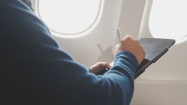 Genç adam uçakta seyahat ederken tablet bilgisayar kullanıyor.