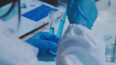 Yakın plan. Mavi takım elbiseli koruyucu eldivenli bir bilim adamının elinde deney tüpü var ve laboratuvara bakıyor. Coronavirus konsepti.