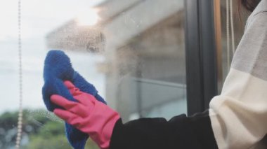 Genç Asyalı kadın modern ofiste sprey ve bezle pencereyi temizliyor.