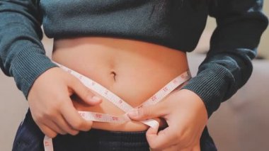 Genç Asyalı kadın, beyaz arka planda ölçüm bandıyla göbeğini ölçüyor. Sağlıklı ve sağlıksız beslenme kavramı.