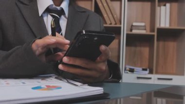 Asyalı profesyonel, parlak ve modern bir ofiste akıllı telefon kullanarak finansal raporlar ve grafikler üzerinde çalışıyor..