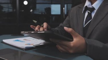 Asyalı işadamı, modern bir ofis ortamındaki tablet üzerindeki mali belgeleri ve grafikleri analiz ediyor.