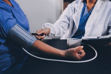 Hastanın tansiyonunu ölçen bir doktor. Hastanın kolunda tansiyon aleti var. Doktor da tansiyon monitörü kullanıyor..