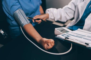 Hastanın tansiyonunu ölçen bir doktor. Hastanın kolunda tansiyon aleti var. Doktor da tansiyon monitörü kullanıyor..