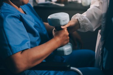 Evde dambıl kullanarak fizyoterapist eğitimi alan yaşlı bir kadın. Terapist, huzurevi egzersizlerinde yaşlı kadına yardım ediyor. Yaşlı bir hasta kollarını uzatmış halterler kullanıyor..