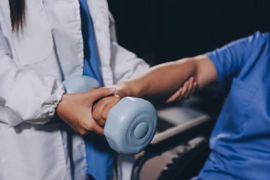 Evde dambıl kullanarak fizyoterapist eğitimi alan yaşlı bir kadın. Terapist, huzurevi egzersizlerinde yaşlı kadına yardım ediyor. Yaşlı bir hasta kollarını uzatmış halterler kullanıyor..