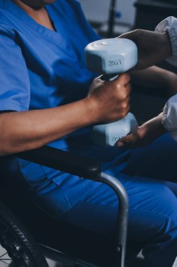 Evde dambıl kullanarak fizyoterapist eğitimi alan yaşlı bir kadın. Terapist, huzurevi egzersizlerinde yaşlı kadına yardım ediyor. Yaşlı bir hasta kollarını uzatmış halterler kullanıyor..