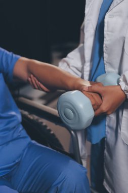 Evde dambıl kullanarak fizyoterapist eğitimi alan yaşlı bir kadın. Terapist, huzurevi egzersizlerinde yaşlı kadına yardım ediyor. Yaşlı bir hasta kollarını uzatmış halterler kullanıyor..